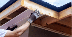 Step-by-step photo of homeowner inspecting fascia board for rot by pressing on wood under roof overhang.